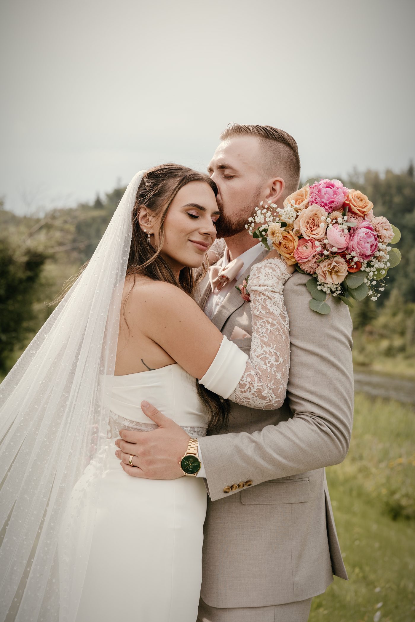 The bride, eyes closed, held against her groom, a bouquet of peach and pink roses at his shoulder