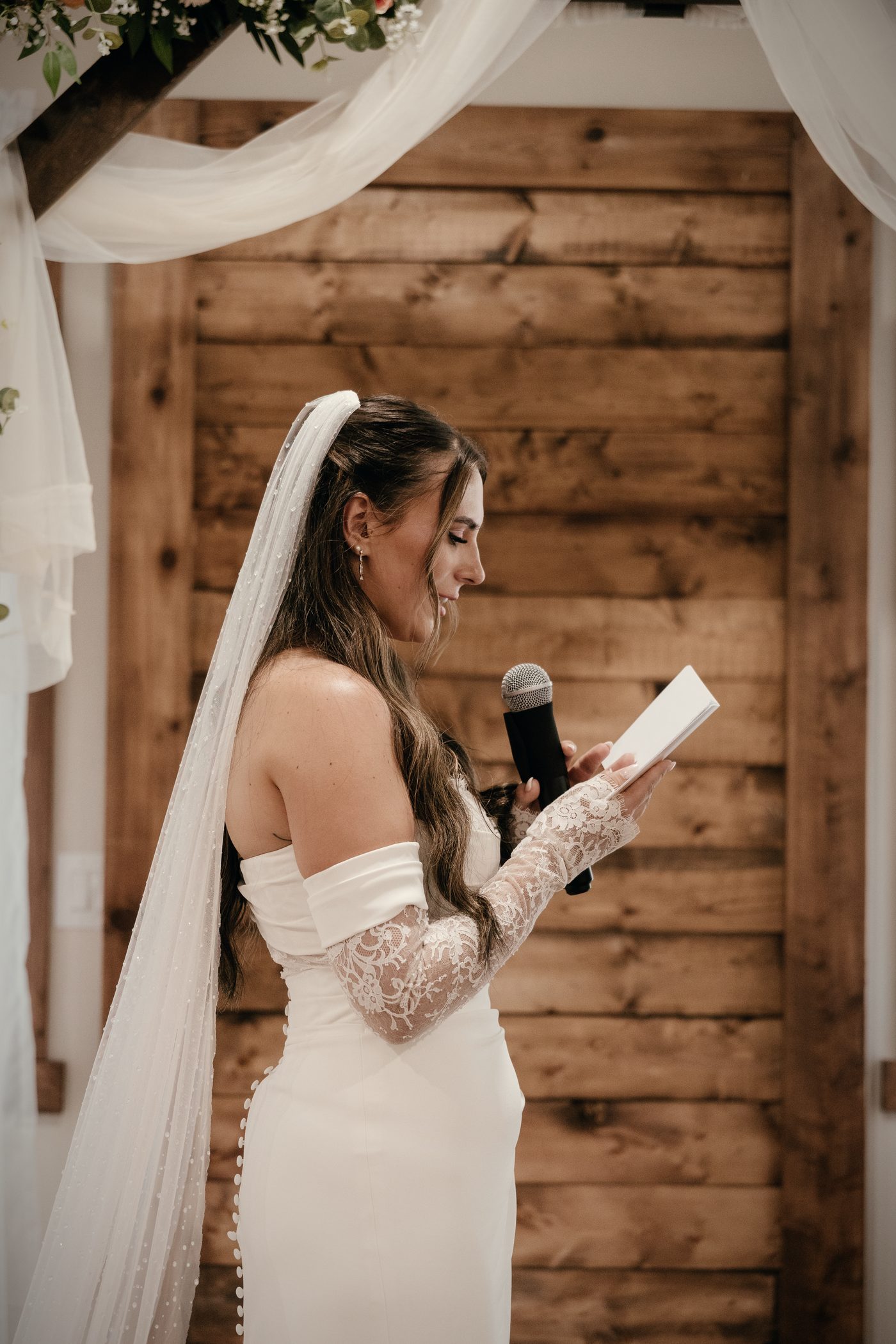 The bride in profile reading her vows, veil falling down her back, the wood wall behind