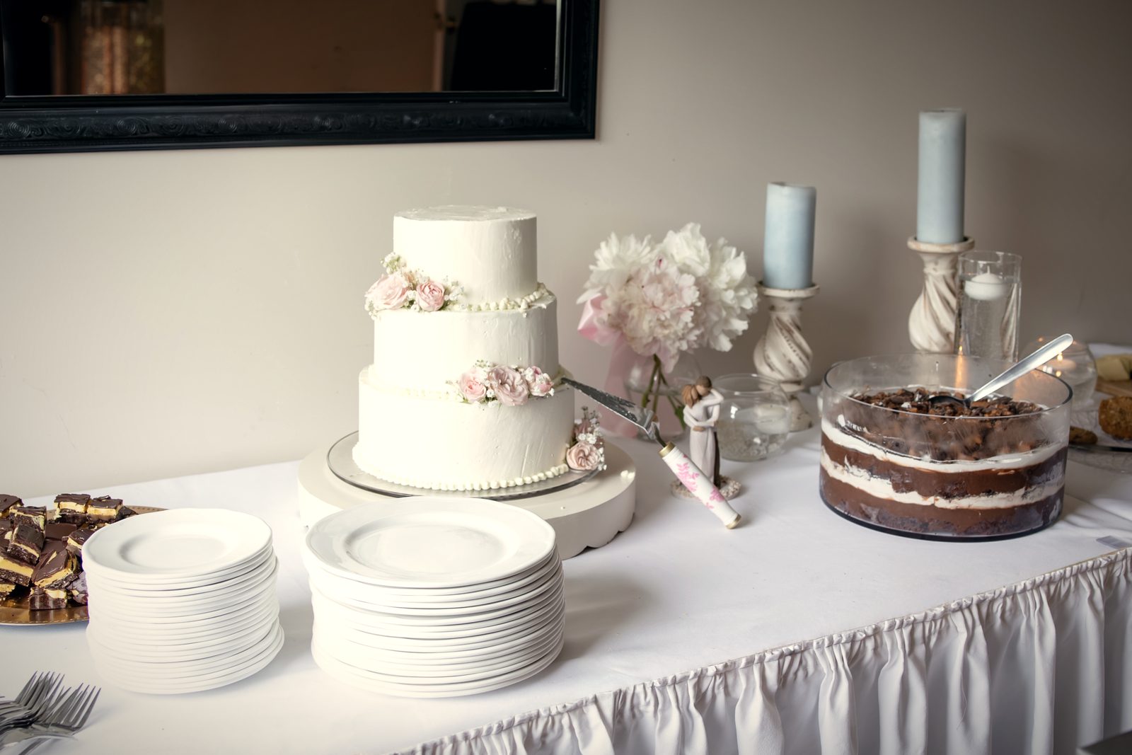 A three-tier cake with fresh florals beside a naked cake and stacked plates on the dessert table