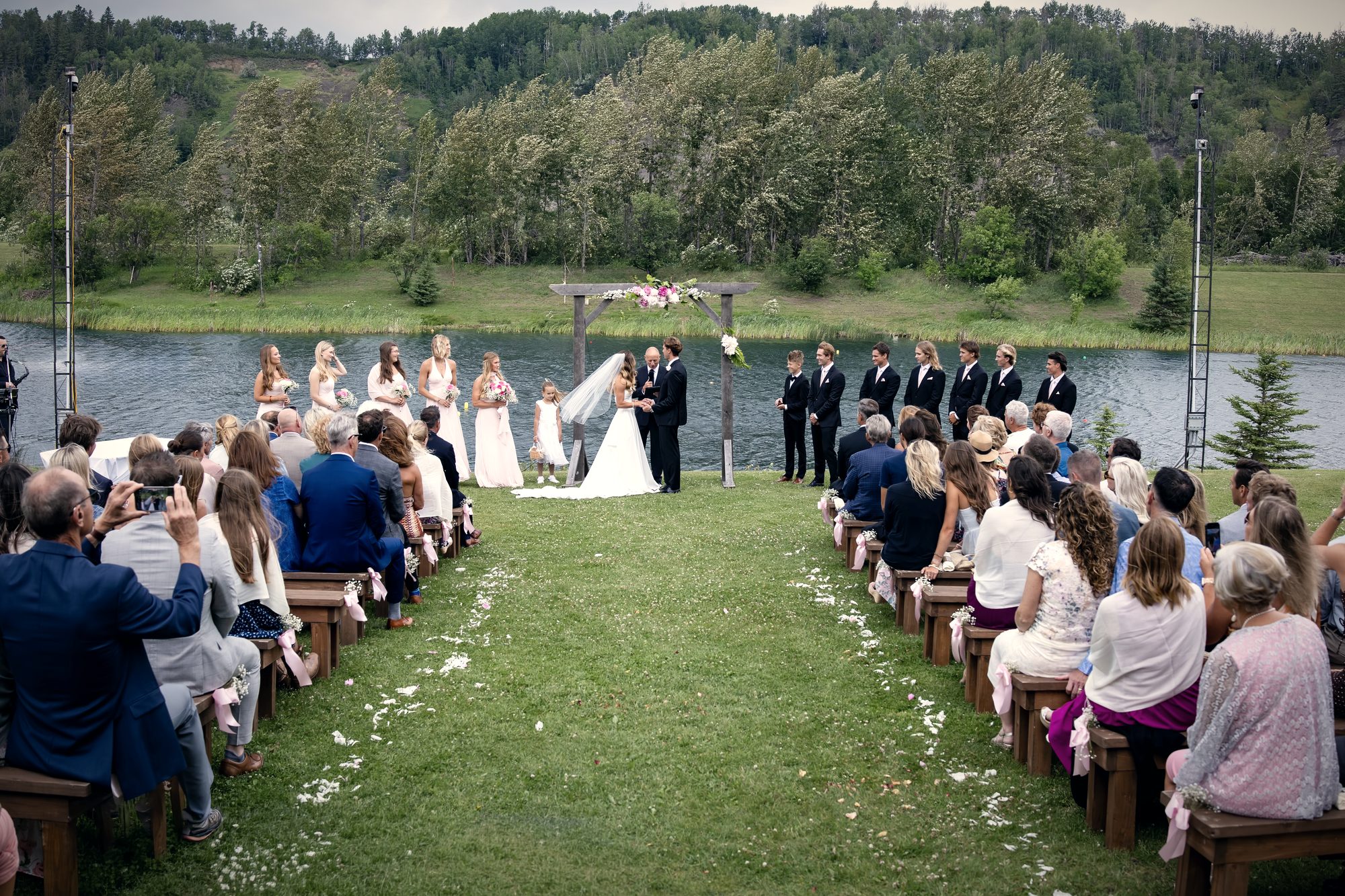 A ceremony at the arbour by the water, guests on wooden benches either side of a petal-strewn aisle