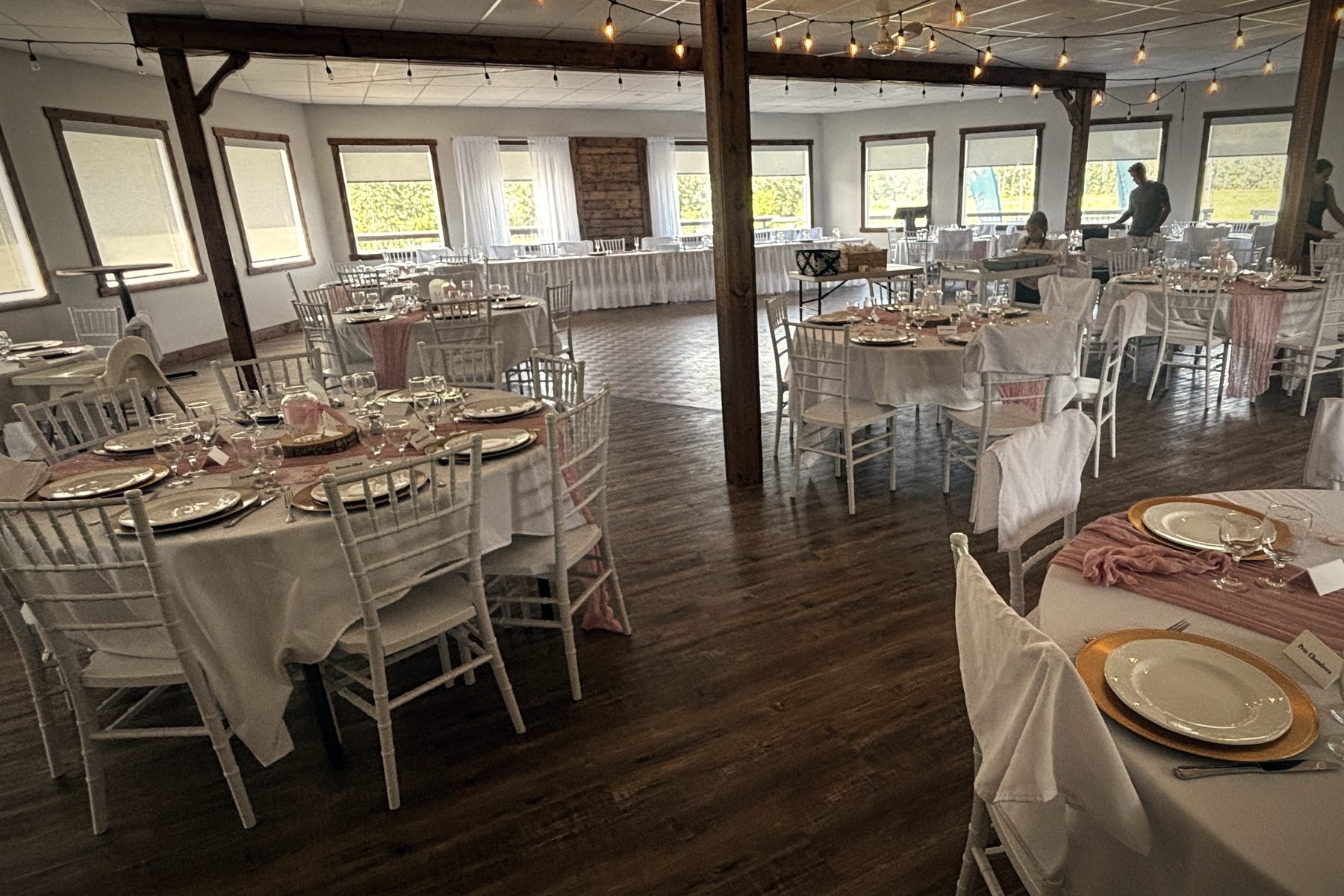 Round tables set for dinner on the hall's wood floor, white chairs, gold chargers and blush napkins