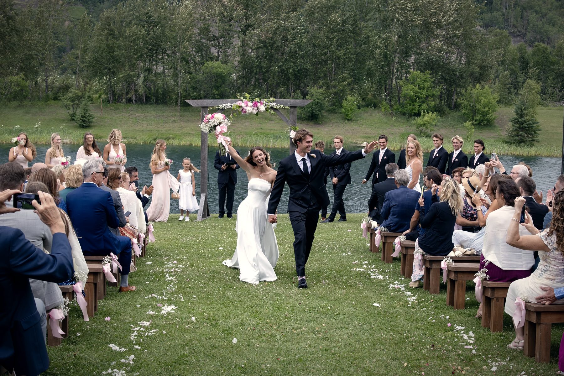 The couple walking back up the aisle through thrown petals, guests cheering from the benches