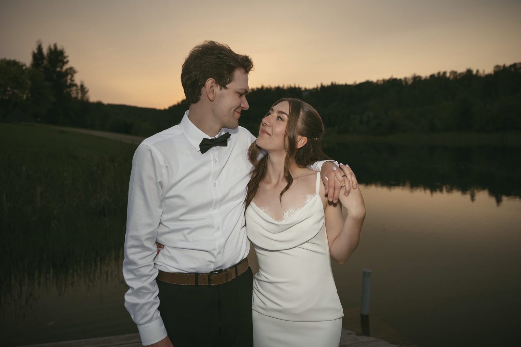 A couple at the water's edge at dusk, the sky still orange behind the treeline