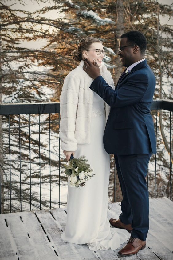 A groom touching his bride's face on a snowy deck, evergreens behind them