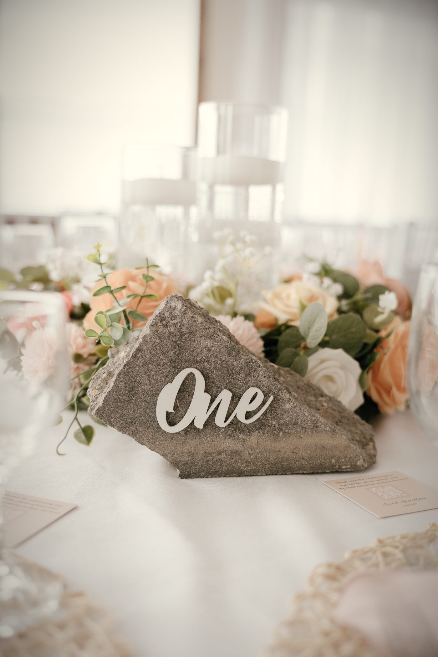 A stone table number reading One among peach roses, white blooms and eucalyptus, a pink napkin beside it