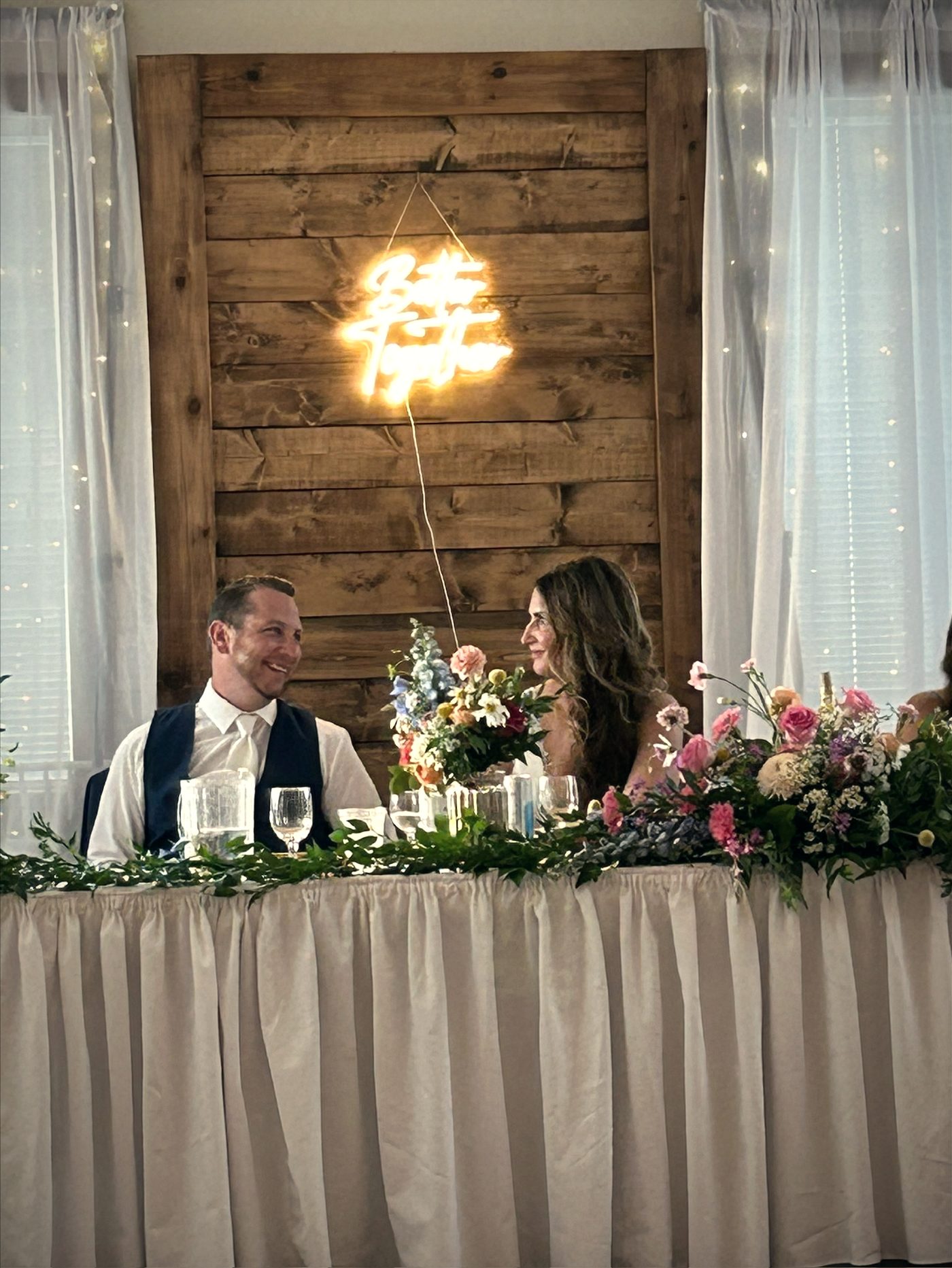 A couple at the head table in front of the wood feature wall, smiling at one another over spring flowers