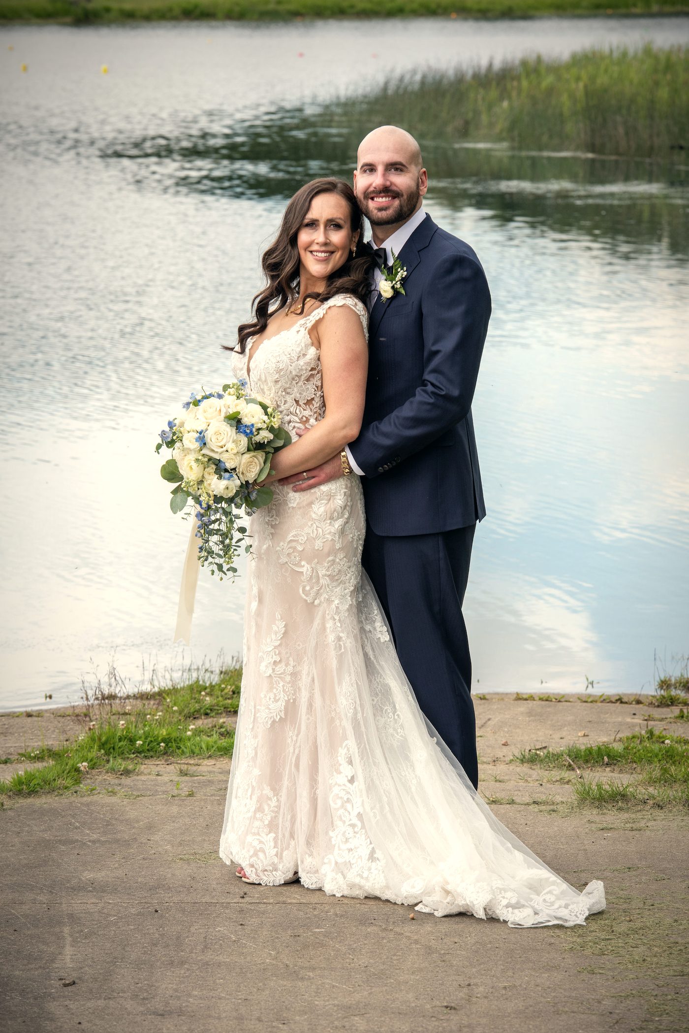 A bride and groom holding each other on the shore, the lake bright behind them
