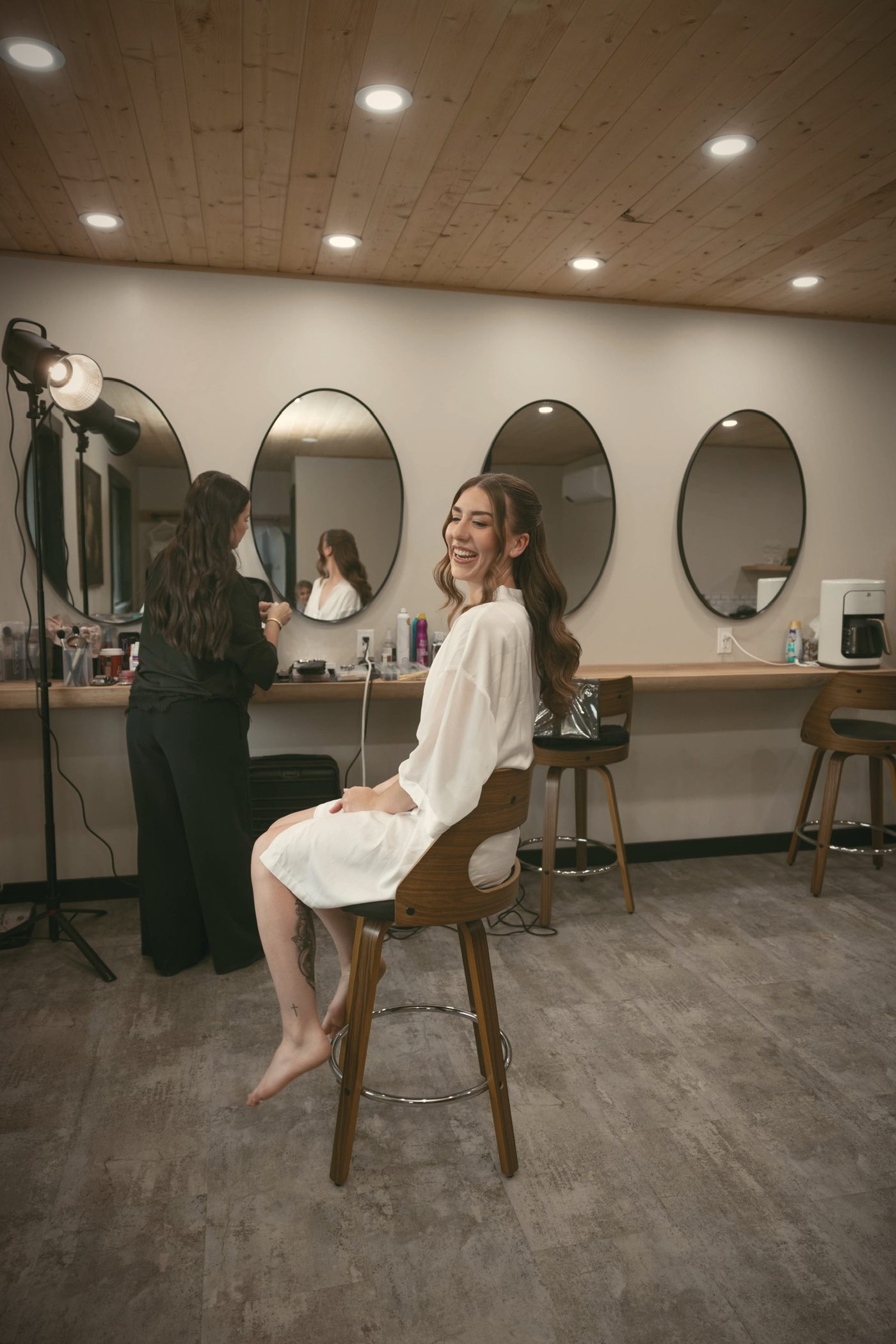 A bride in a robe laughing in the suite, a ring of round mirrors above the make-up counter