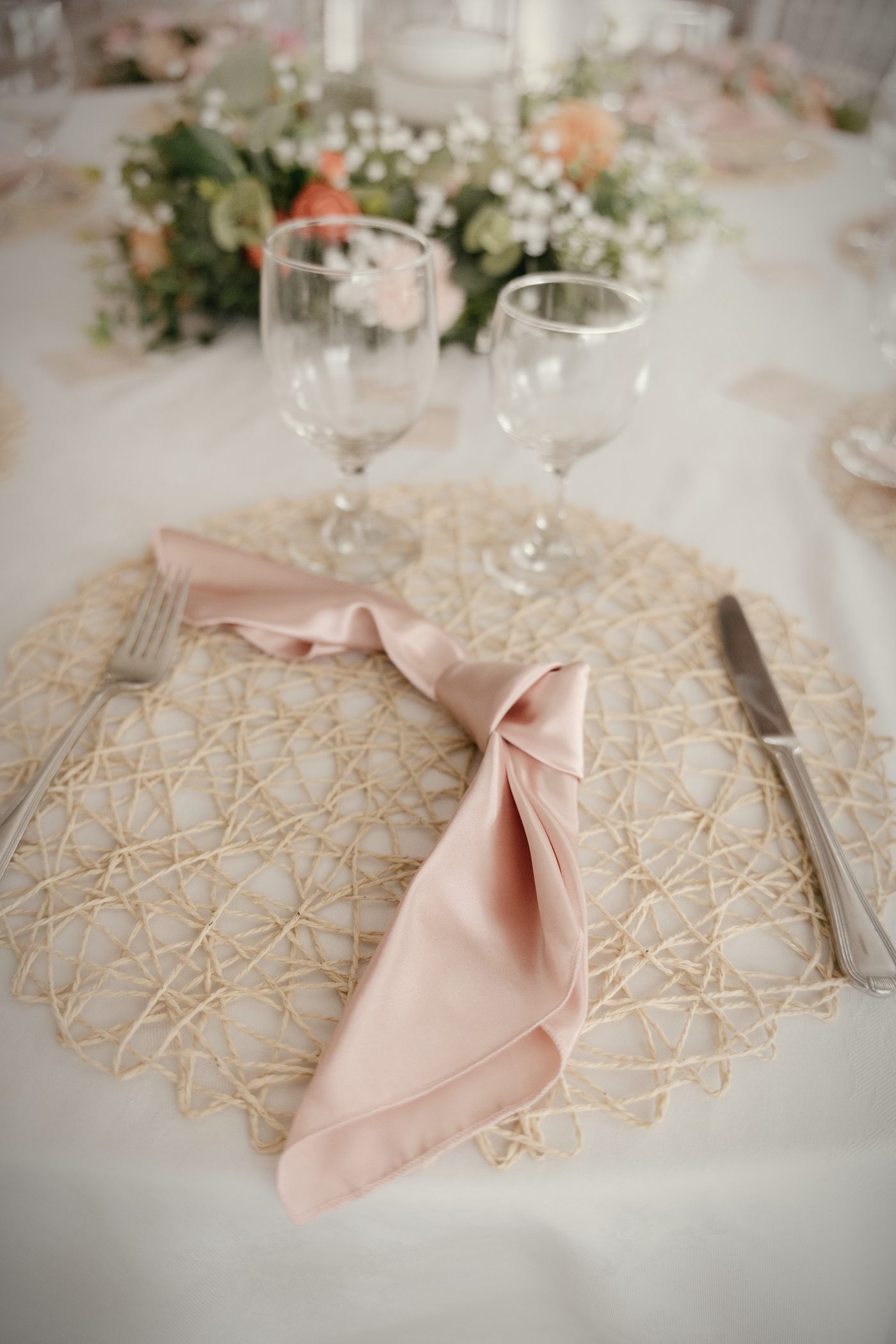 A blush napkin knotted on a woven charger, florals soft behind it