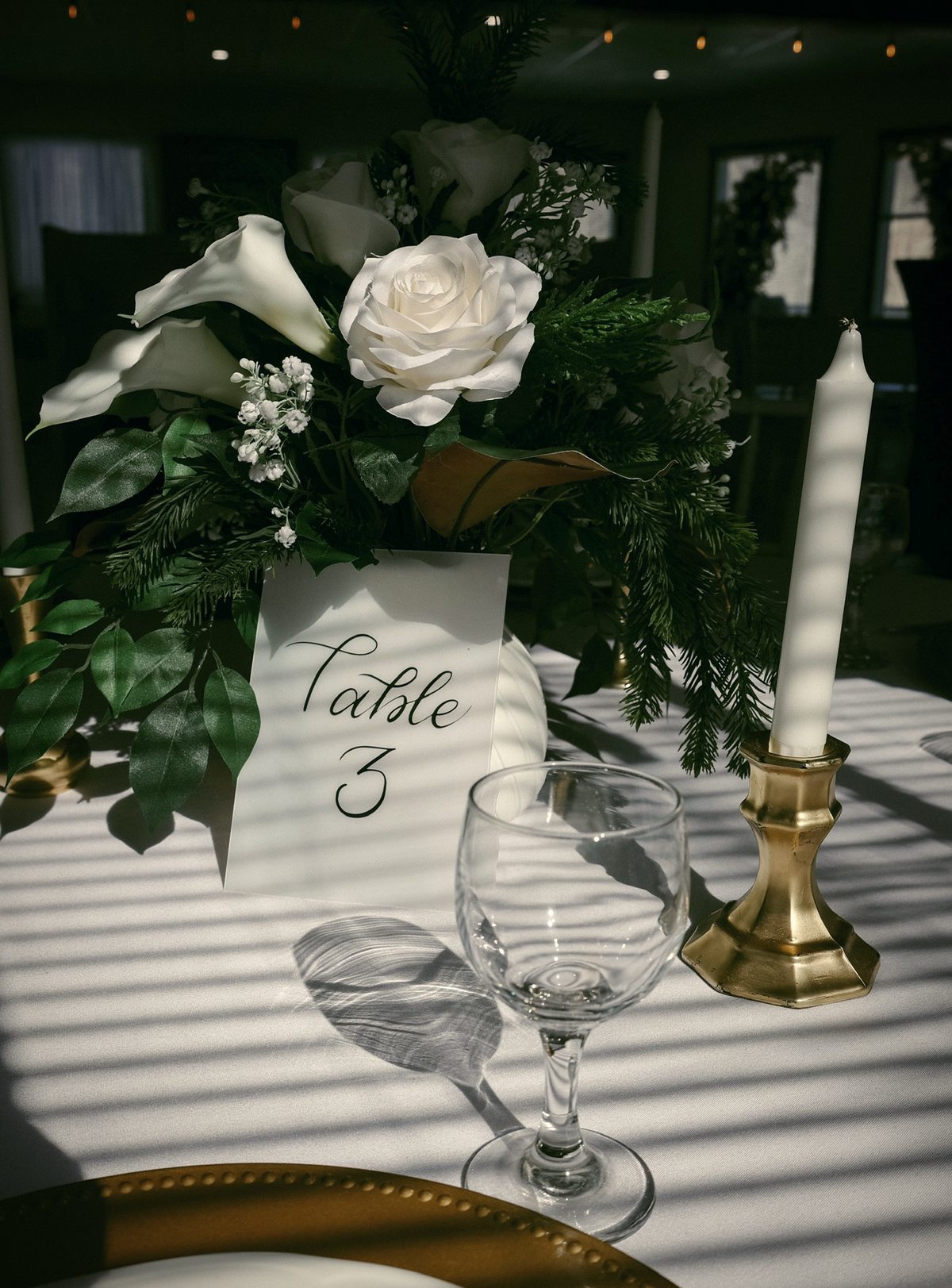A table number in script beside white roses, a candle and gold-rimmed plates