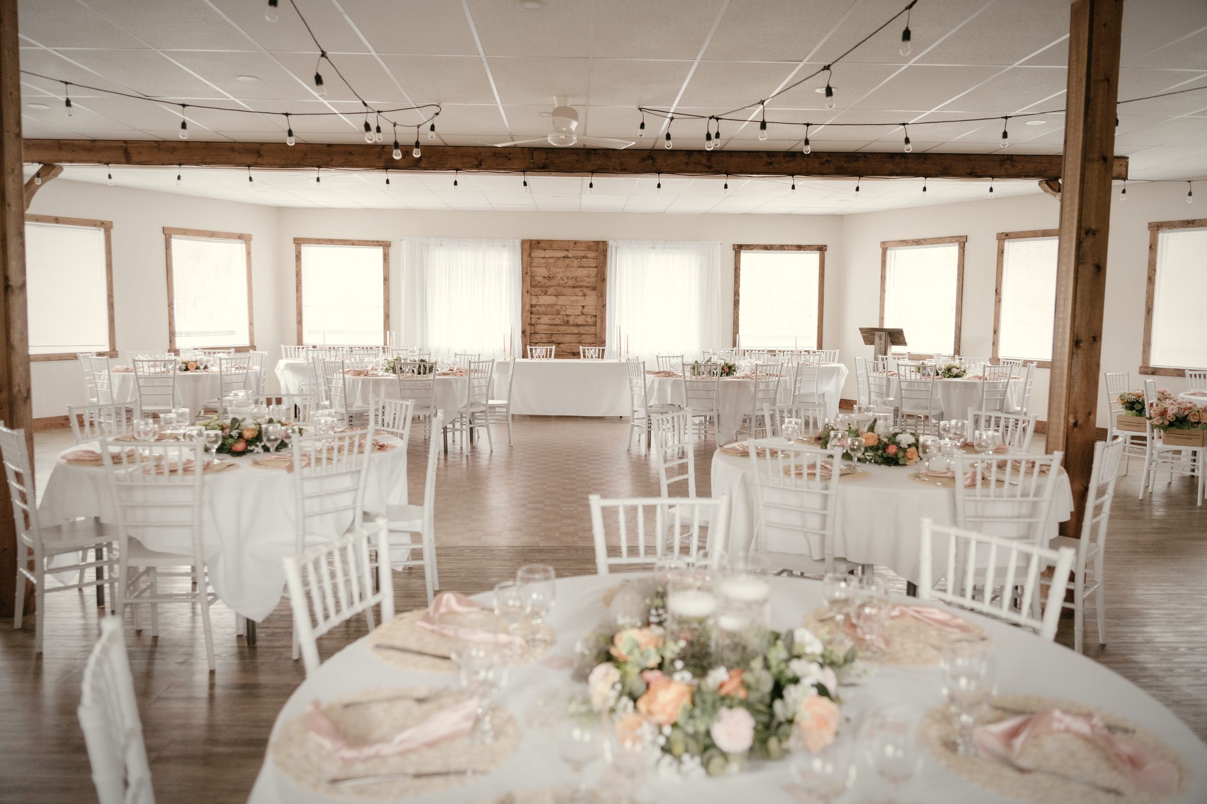 The hall set for dinner: round tables in white linen with low florals, timber posts and a beam, string lights overhead and tall windows along the far wall
