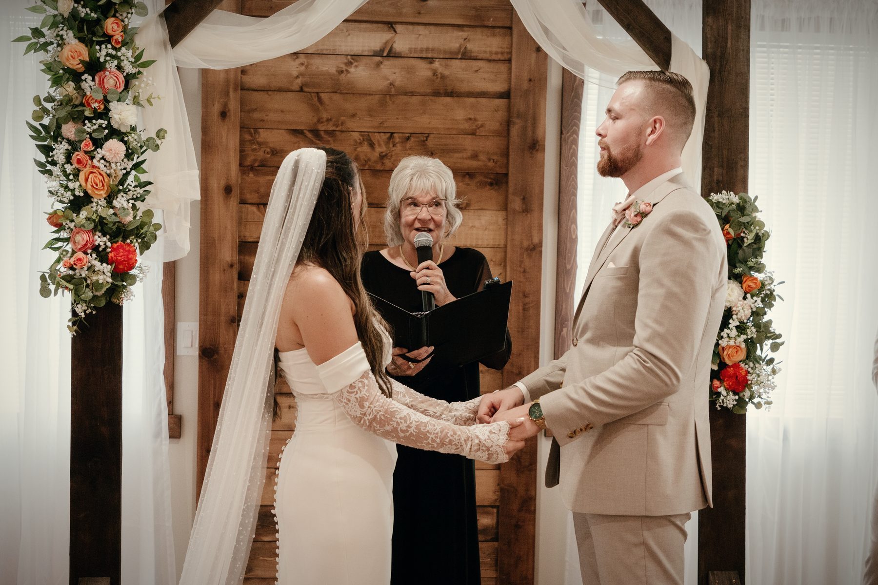 Hands joined at the timber arch in the hall, the officiant between the couple, white drapery and florals behind them