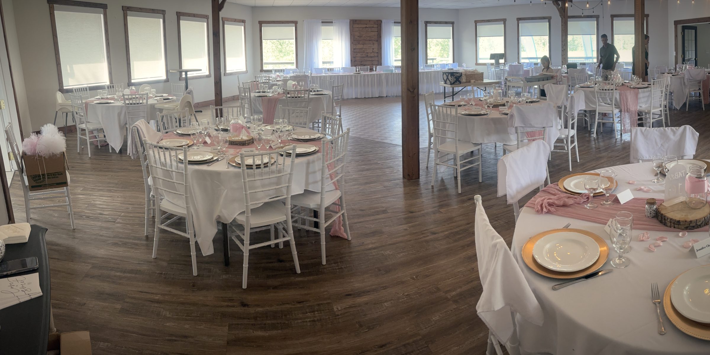 The hall set for dinner by day: round tables in white linen with blush napkins and gold chargers, timber posts, string lights and windows down both sides