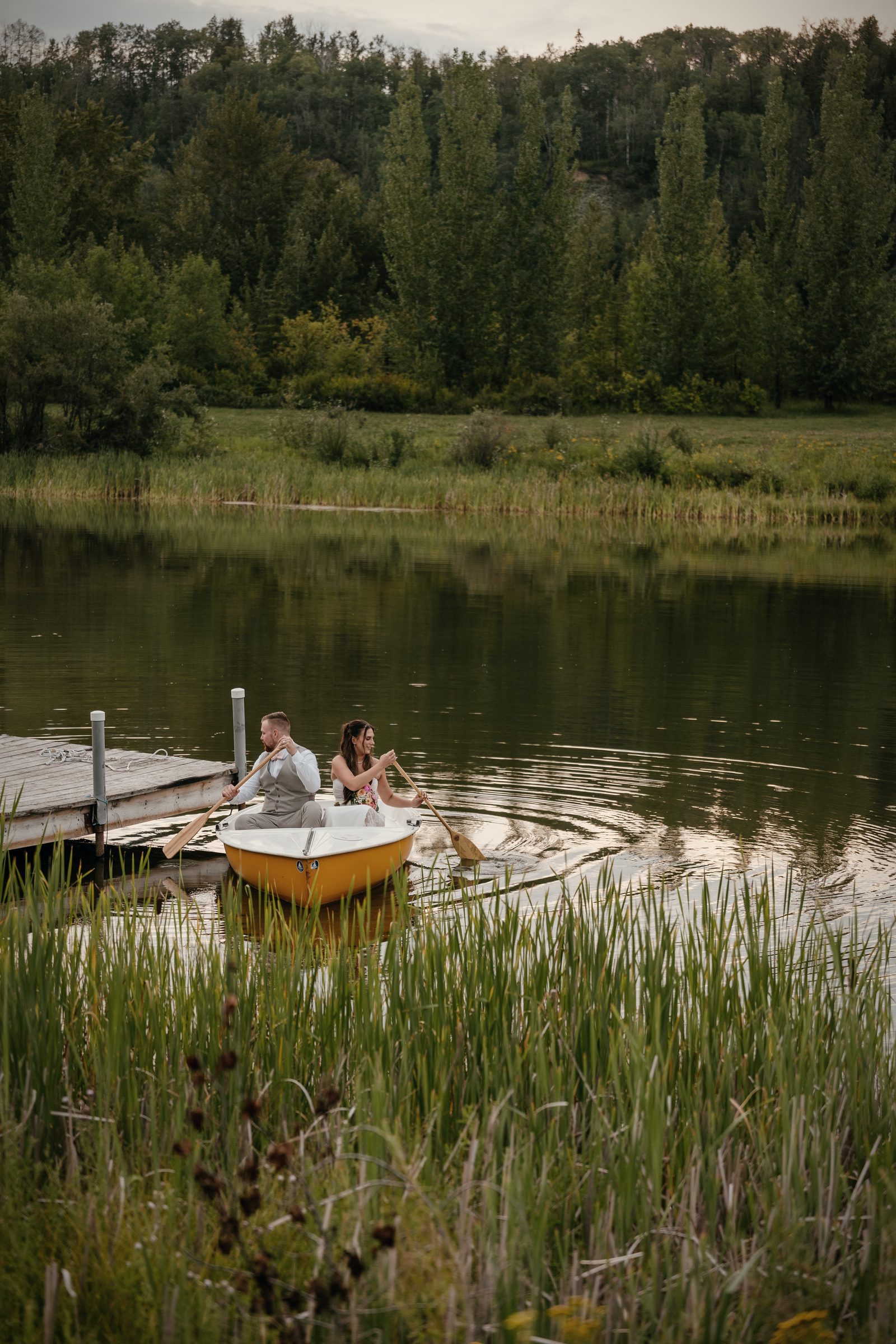 A couple on the dock with the yellow rowboat tied beside them, reeds in the foreground and the lake all around