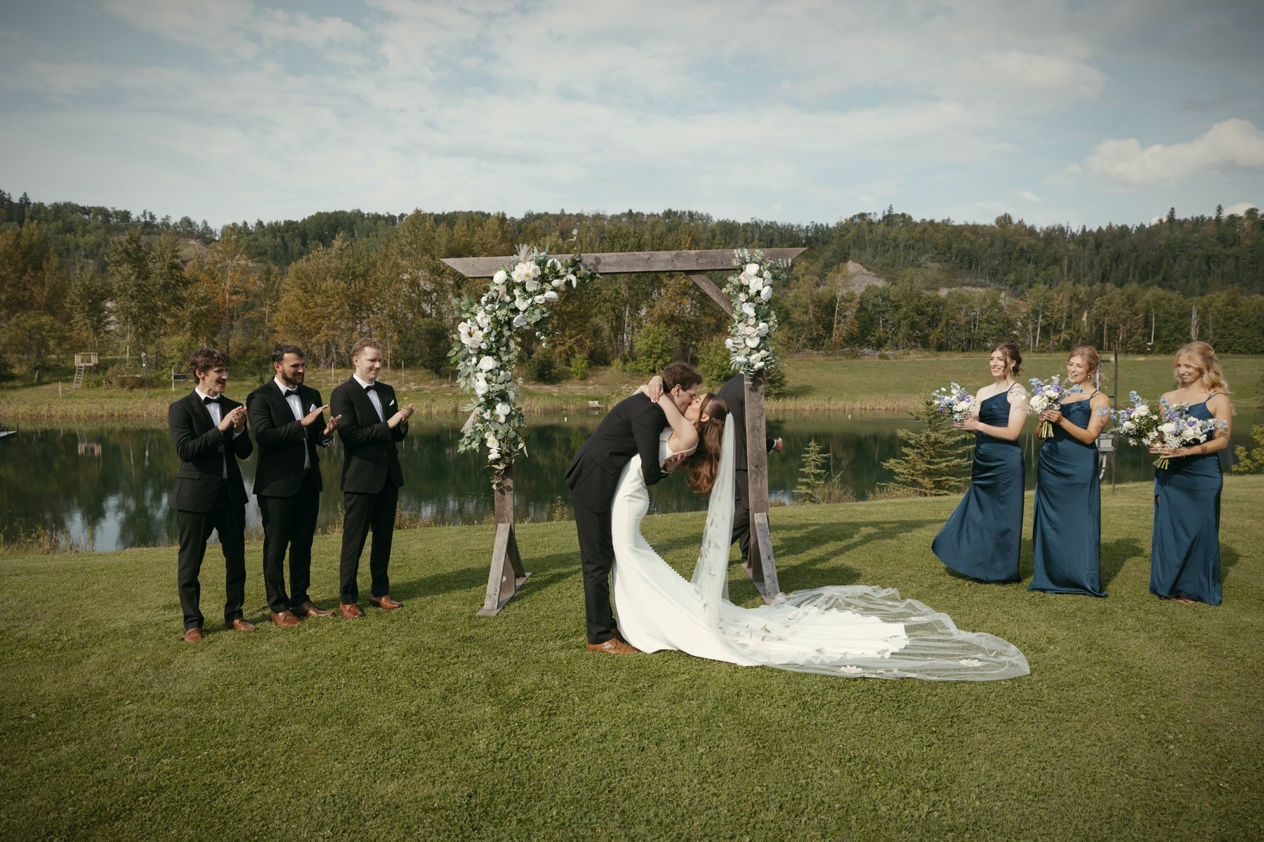 The couple kissing at the flowered arbour, the wedding party either side and the lake behind