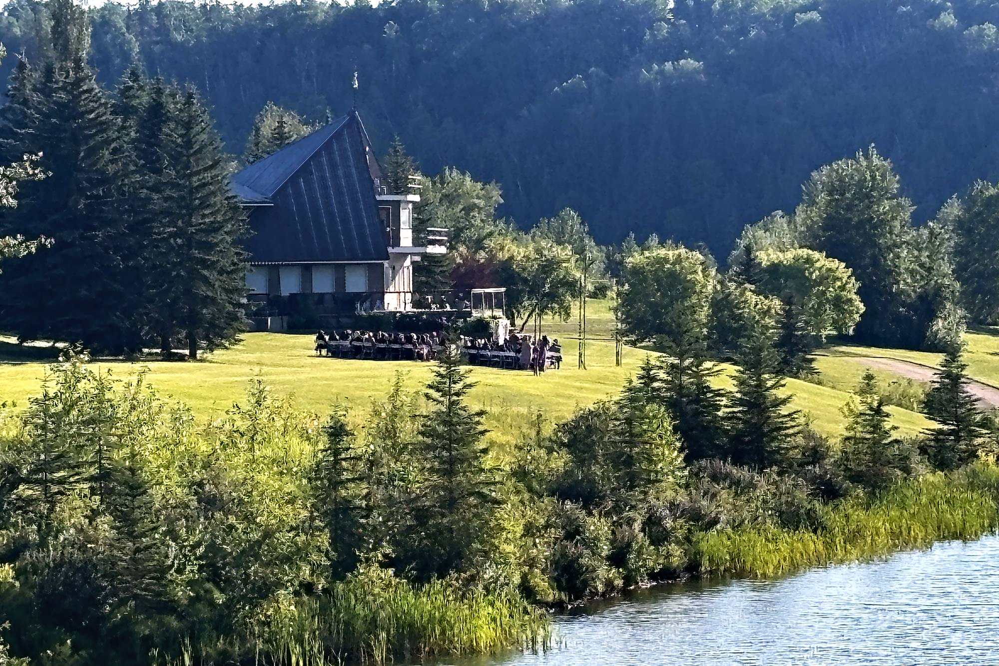 The hall on the slope above the water, seen across the lake through spruce