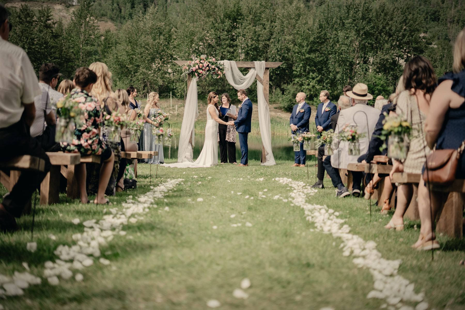 A ceremony on the lawn, guests on wooden benches either side of a petal-strewn aisle